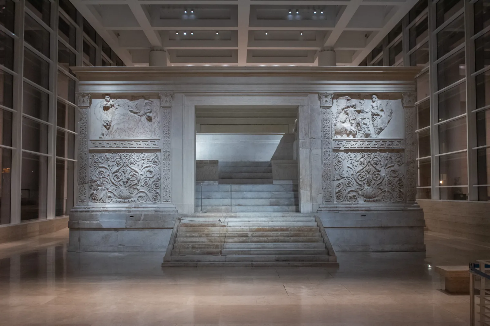 A rectangular marble enclosure carved with processional figures above a deep band of acanthus scrollwork, standing inside a modern glass-walled hall.