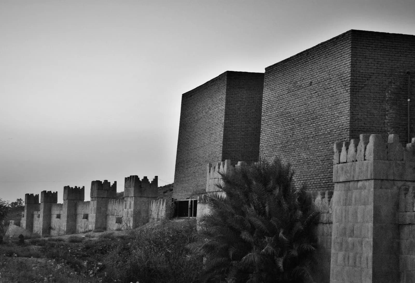 A rebuilt Assyrian city gate of pale mudbrick, with a high arched opening flanked by stepped towers, standing on a low mound.