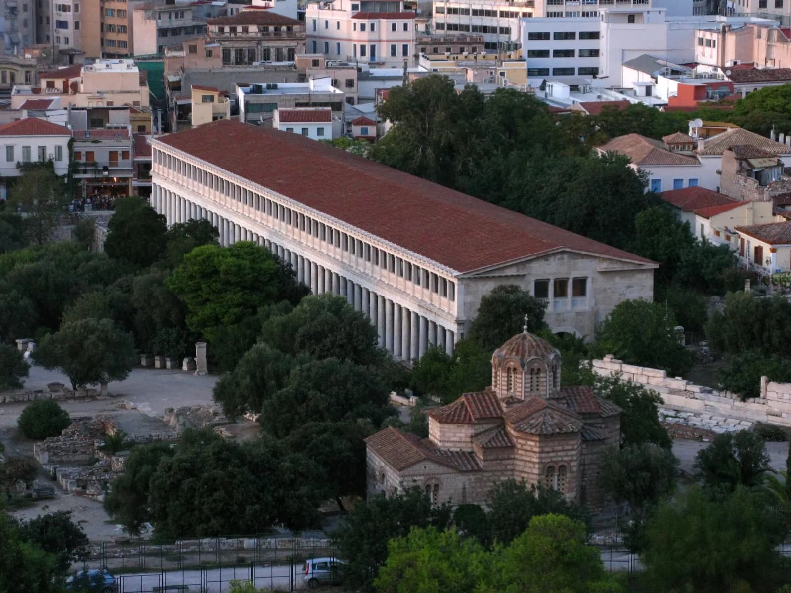 A long two-storey colonnaded building with a tiled roof, complete and freshly built in appearance, standing at the edge of an excavated square.