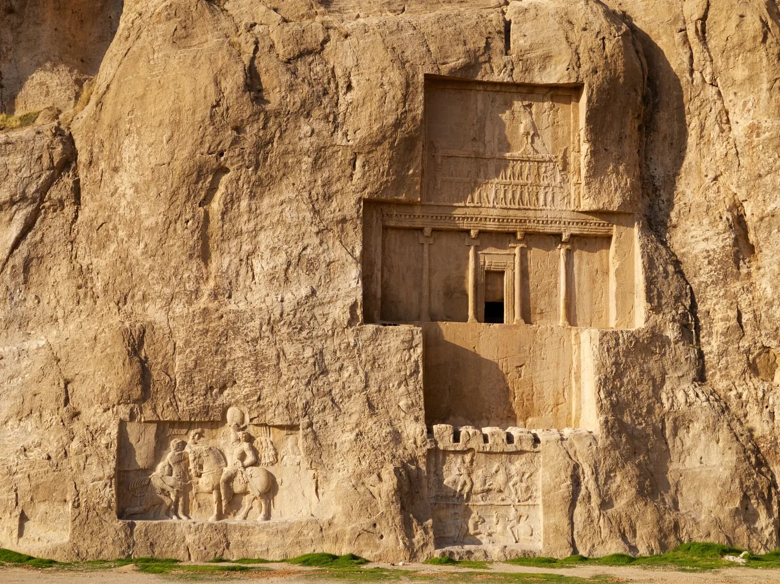 A cross-shaped façade carved high into a cliff, with a blind columned porch below and a relief of a figure on a platform above, and a separate later relief cut into the rock beneath.