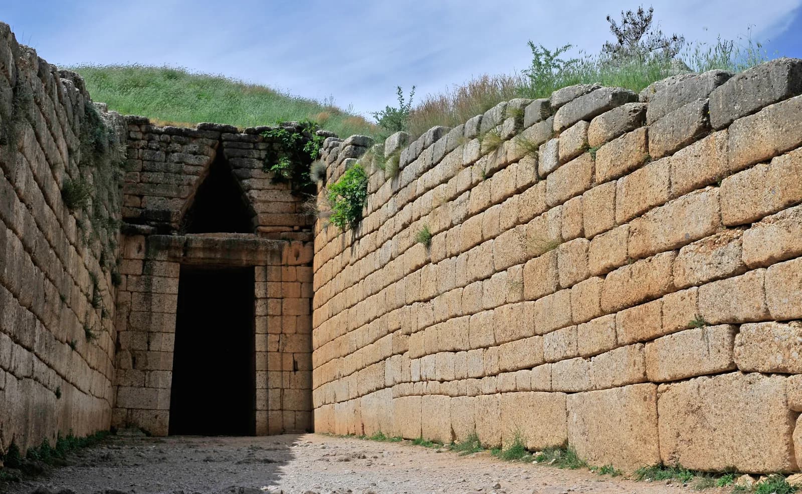 A long walled passage cut into a hillside leading to a tall stone doorway with a massive lintel and a triangular opening above it.