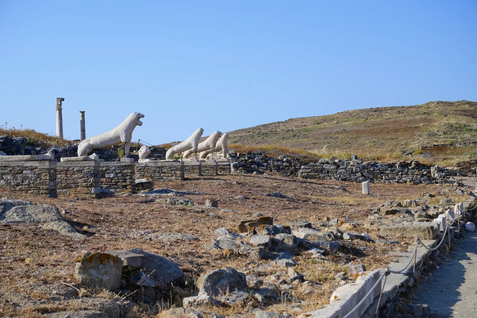 A row of weathered marble lions on a low terrace, seated on their haunches with heads turned, above scattered foundations on a bare island.
