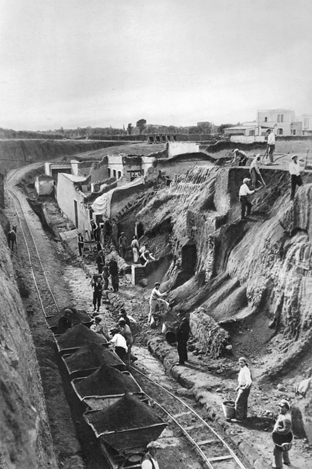 A historical photograph of a large excavation in progress, with workmen, spoil baskets and a light railway among partly cleared Roman walls.